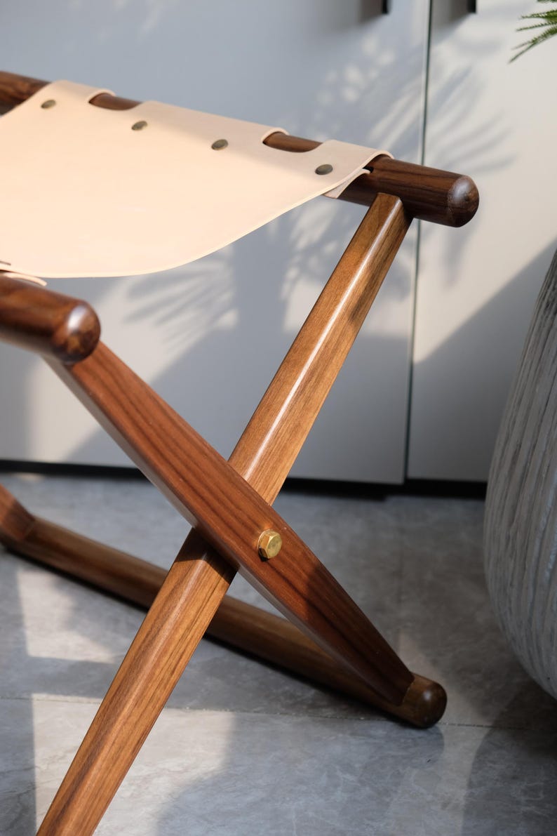 May include: A wooden folding chair with a light brown leather seat. The chair has a simple design with a cross-shaped frame and rounded legs.