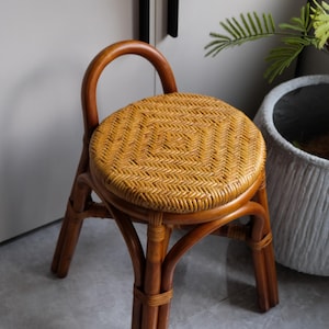 May include: A brown wicker stool with a woven seat and a rounded back. The stool is sitting on a gray tiled floor.
