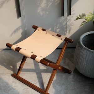 May include: A wooden folding stool with a light brown leather seat. The stool has a simple design with a cross-shaped frame and rounded legs.