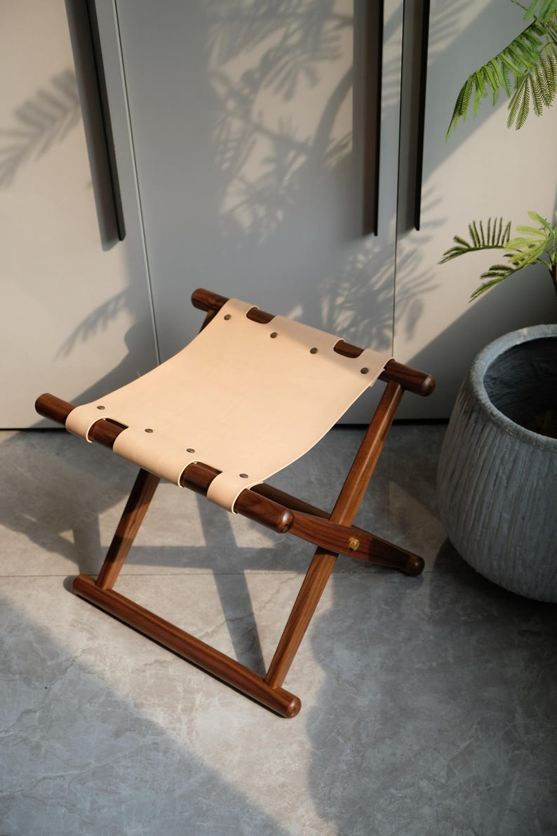 May include: A wooden folding stool with a light brown leather seat. The stool has a simple design with a cross-shaped frame and rounded legs.