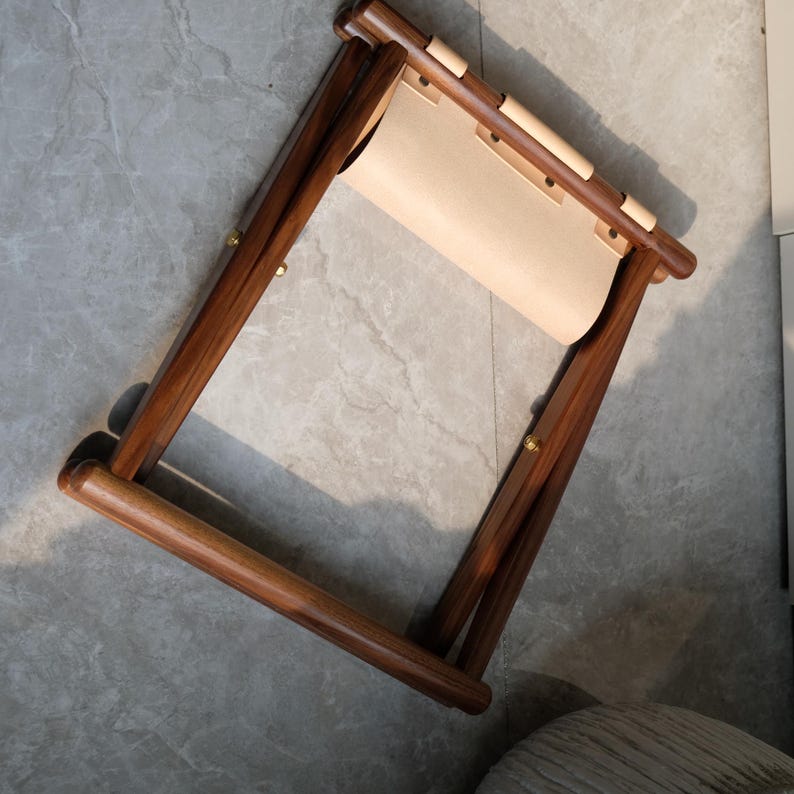 May include: A wooden folding stool with a light brown leather seat. The stool is made of dark brown wood and has a simple design.