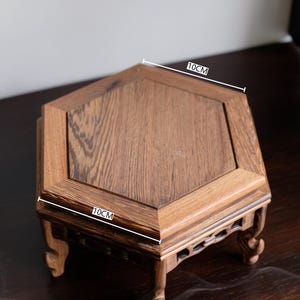 May include: Antique hexagonal wooden stand with a dark brown finish. The stand measures 10 inches in diameter and has intricate carvings on the legs.