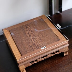 May include: A square wooden stand with a 23-inch by 23-inch top and carved legs. The stand is made of dark wood and has a natural finish.