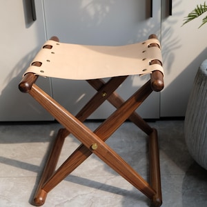 May include: A wooden folding stool with a light brown leather seat. The stool has a simple, classic design and is perfect for adding extra seating to any room.
