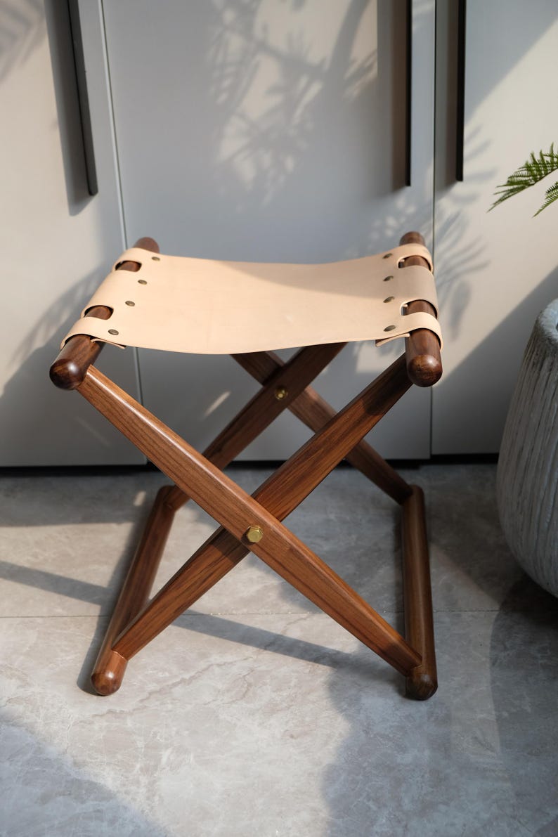 May include: A wooden folding stool with a light brown leather seat. The stool has a simple, classic design and is perfect for adding extra seating to any room.
