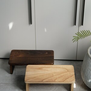 May include: Two small wooden step stools. One is a dark brown color, and the other is a light natural wood color. Both have a rectangular top and four short legs. The stools are placed on a gray tiled floor.