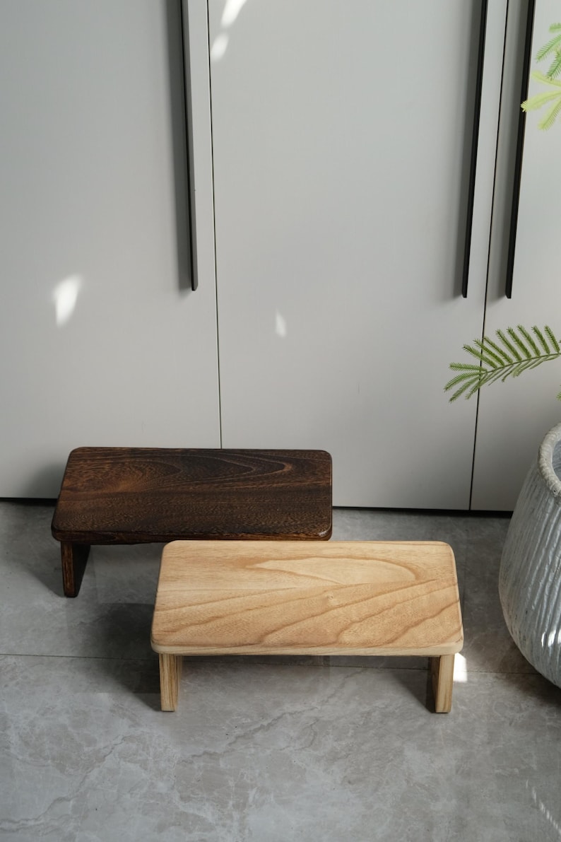 May include: Two small wooden step stools. One is a dark brown color, and the other is a light natural wood color. Both have a rectangular top and four short legs. The stools are placed on a gray tiled floor.