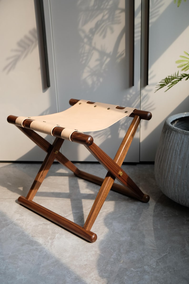 May include: A wooden folding stool with a light brown leather seat. The stool has a simple, minimalist design and is made of dark wood.