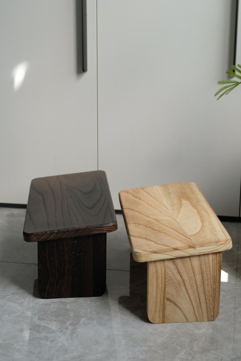 May include: Two wooden stools, one dark brown and one light brown, are displayed side-by-side. Each stool has a rectangular seat and a square base. The stools are made of wood and have a natural grain.