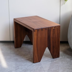 Puede incluir: Taburete de madera artesanal con tapa rectangular y patas anguladas. Fabricado en madera marrón oscura, con uniones visibles en las esquinas. Diseño moderno y minimalista, adecuado para diversos interiores. Mide unos 40 cm de largo.