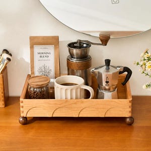May include: A rectangular wooden tray with a scalloped front edge and four spherical feet. It holds a brown paper bag labelled Morning Blend, a glass jar with a wooden lid, a white ribbed ceramic mug, a manual coffee grinder, and a metal moka pot.