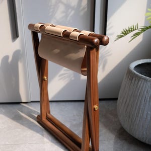May include: A wooden folding stool with a light brown leather seat. The stool is made of dark brown wood and has a brass accent.