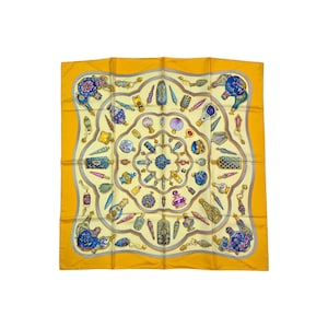 May include: A square silk scarf with a yellow background and a repeating pattern of perfume bottles, shells, and decorative elements in blue, gold, and cream. The scarf has a gray border.