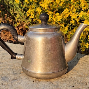 以下が含まれることがあります： 黒い取っ手と蓋に黒いノブが付いた銀色のティーポット。ティーポットは灰色の表面の上に置かれています。