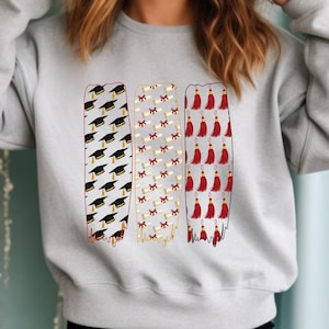 May include: A light gray sweatshirt featuring three vertical columns with graduation-themed designs. The columns display graduation caps, diplomas, and red tassels. The designs are outlined in black and red.