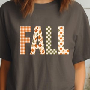 May include: A charcoal grey t-shirt featuring the word FALL in large block letters. Each letter has a unique pattern: the F is orange and white gingham, the A has autumn leaves, the L is green and white checkered, and the L has small pumpkins.