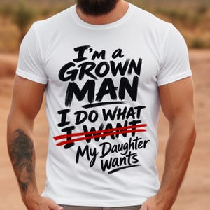 May include: A white short-sleeved t-shirt featuring black text that reads I'm a GROWN MAN I DO WHAT I WANT MY DAUGHTER WANTS. The word WANT is crossed out with two horizontal red lines, creating a bold graphic design on the front of the garment.