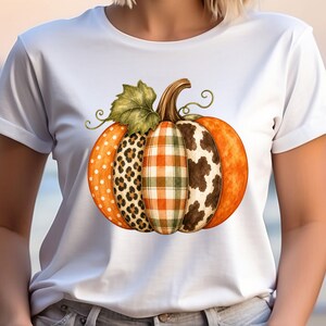 May include: A white short-sleeved t-shirt featuring a large central graphic of a pumpkin. The pumpkin is divided into vertical segments with different patterns, including orange polka dots, leopard print, plaid, cow print, and solid orange.