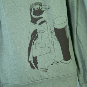 May include: A green long-sleeved sweatshirt with a black and white illustration of a penguin wearing a vest and holding a glass of beer.