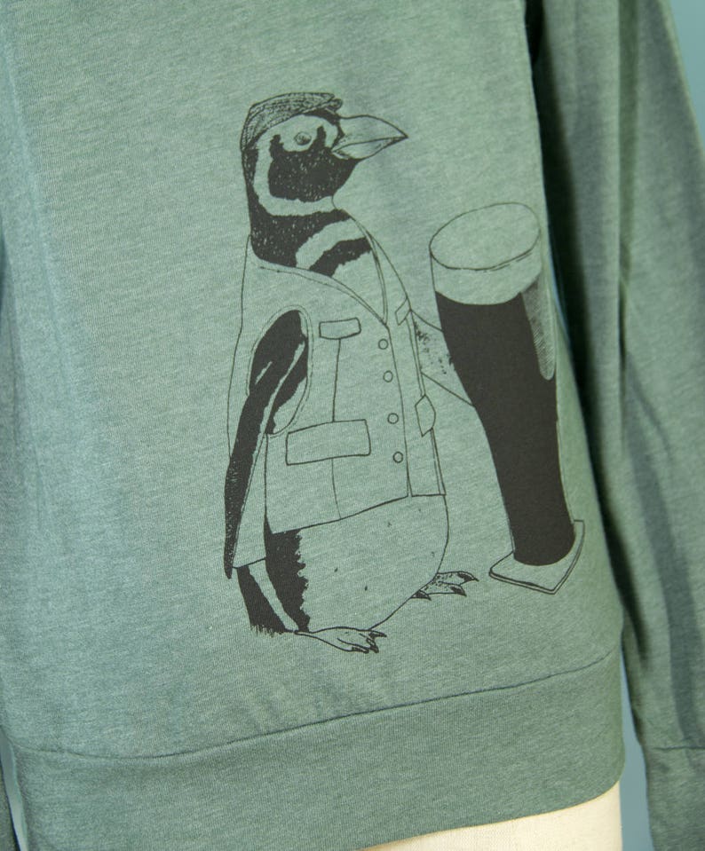 May include: A green long-sleeved sweatshirt with a black and white illustration of a penguin wearing a vest and holding a glass of beer.