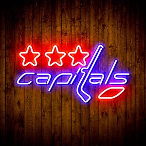Op de afbeelding: Neonbord met het woord "capitals" in blauw en wit, met drie rode sterren erboven. Een hockeystick en een rode ovaal zijn ook in het ontwerp opgenomen. De achtergrond is van hout.