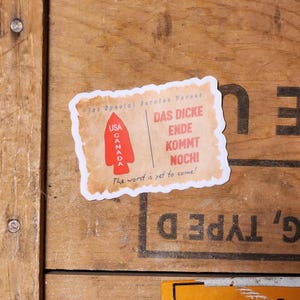 May include: A white sticker with a red arrow pointing down. The arrow is labelled "USA" and "CANADA". The sticker also has the text "DAS DICKE ENDE KOMMT NOCHI" and "The worst is yet to come!"