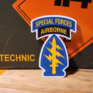 May include: A white sticker with a blue and yellow Special Forces Airborne emblem. The emblem features a lightning bolt and a sword inside a blue arrowhead.