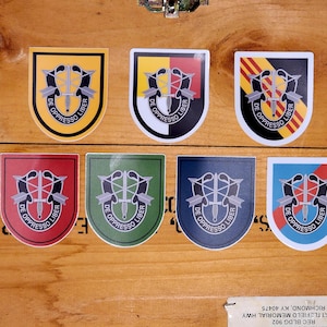 Op de afbeelding: Zeven verschillende gekleurde vinyl stickers met hetzelfde ontwerp. Het ontwerp is een gestileerde zwart-wit afbeelding van een dolk met twee pijlen die erachter gekruist zijn. De tekst "DE OPPRESSO LIBER" staat in een cirkel rond de dolk. De stickers zijn geel, rood, groen, blauw, zwart en twee verschillende tinten geel.