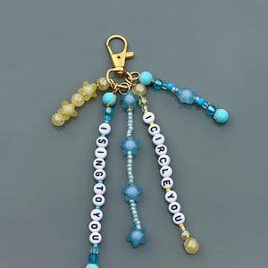 May include: A gold-toned lobster clasp keychain featuring multiple dangling strands of beads. Two strands contain white letter beads spelling out I SING TO YOU and I CIRCLE YOU. Other strands feature blue and yellow star-shaped beads and small round beads.