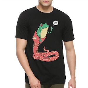 Op de afbeelding: Zwart T-shirt met een afbeelding van een groene kikker die op een rode slang rijdt met het woord "OK" in een spraakballon boven de kikker.