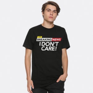 以下が含まれることがあります： 「LIVE BREAKING NEWS I DON'T CARE!」という白いグラフィックがプリントされた黒いTシャツ
