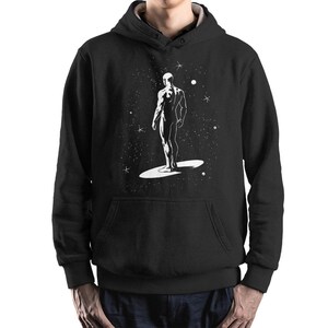 May include: Black hoodie with a white graphic depicting a figure on a surfboard-like shape against a starry backdrop. The design is a minimalist illustration with a cosmic theme, suitable for those interested in space and art.