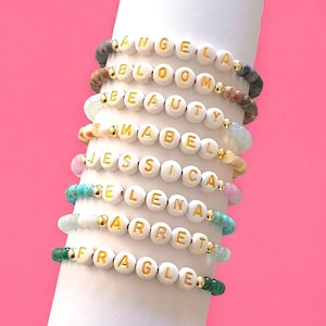 May include: A stack of eight bracelets with different colored beads and white beads with gold lettering spelling out names, including "Angela", "Bloom", "Beauty", "Mabel", "Jessica", "Elena", "Arret", and "Fragile".