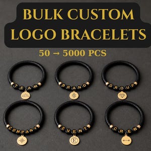 Op de afbeelding: Zwarte leren armbanden met goudkleurige kralen die woorden als "TEAM", "BRAND" en "GROUP" spellen. Elke armband heeft een gouden bedel met een uniek ontwerp. De tekst bovenaan luidt "BULK CUSTOM LOGO BRACELETS".