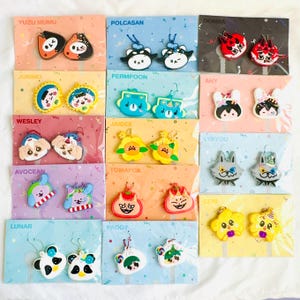 Puede incluir: Una colección de pendientes coloridos de estilo caricatura, presentados en tarjetas individuales. Cada par presenta un diseño de personaje único, incluyendo animales y figuras caprichosas. Las tarjetas están etiquetadas con nombres como "YUZU MUMU" y "POLCASAN".