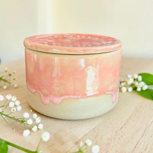Könnte beinhalten: Ein Keramiktopf mit Deckel, mit zweifarbigem Design. Der obere Teil ist glänzend rosa, der in einen cremefarbenen, cremefarbenen Sockel übergeht. Der Topf ist zylindrisch, mit passendem Deckel. Handgefertigtes Stück.