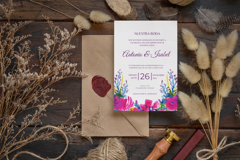 May include: A wedding invitation with a watercolor floral design in shades of pink, blue, and green. The invitation reads "Nuestra Boda" and includes the names Antonio & Isabel, the date Saturday, November 26, 2014, and the time 09 PM. The invitation is on a brown wooden surface with dried flowers and a wax seal.