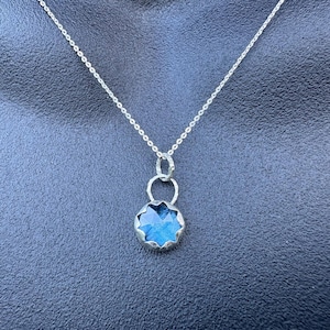 May include: A silver necklace featuring a delicate chain and a round pendant with a faceted blue gemstone. The pendant is secured by a small silver ring and has a scalloped metal edge, showcasing a bright blue centre with a star-like cut.