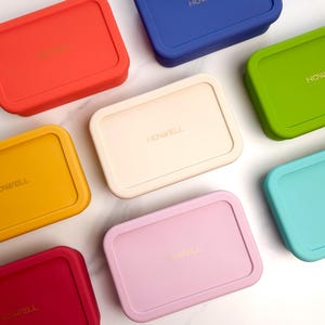 May include: An overhead shot of several rectangular food storage containers in various colors, including red, blue, green, yellow, pink, and cream. Each container has a matching lid and the word "HOWELL" printed on the lid in gold.
