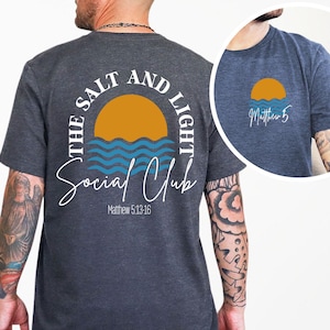 Könnte beinhalten: Dunkelgraues T-Shirt mit einem kreisförmigen Design auf dem Rücken, das eine gelbe Sonne, blaue Wellen und den Text "THE SALT AND LIGHT Social Club" mit "Matthew 5:13-16" darunter zeigt. Eine kleinere Version des Designs befindet sich auf der Vorderseite. Das T-Shirt wird von einer Person getragen.