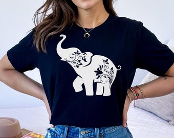 May include: A black t-shirt with a white elephant graphic. The elephant has intricate floral designs on its body. The shirt is being worn by a person with long brown hair.