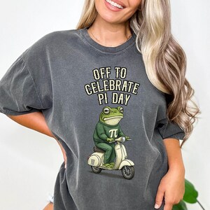 May include: Dark grey t-shirt with the text "OFF TO CELEBRATE PI DAY." The shirt features a cartoon frog wearing a green sweater with a pi symbol, riding a cream-coloured scooter. The frog has a serious expression.