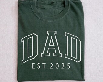 Puede incluir: Una camiseta verde oscuro con texto blanco que dice "DAD EST 2025".