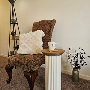 May include: A brown upholstered chair with a white throw pillow sits next to a white column pedestal table with a wooden top. A mug sits on the table with a white coaster. The chair has a brown patterned fabric and wooden legs. The room has beige carpet and white walls.