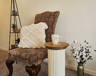 Puede incluir: Una silla tapizada en marrón con un cojín blanco se encuentra junto a una mesa de pedestal de columna blanca con una tapa de madera. Una taza se encuentra sobre la mesa con un posavasos blanco. La silla tiene un tejido marrón estampado y patas de madera. La habitación tiene una alfombra beige y paredes blancas.