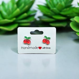 以下が含まれることがあります： 白いカードに「Handmade with Love」と書かれた、チェリーの形をしたイヤリングのペア。イヤリングは赤と緑で、光沢のある仕上げになっています。
