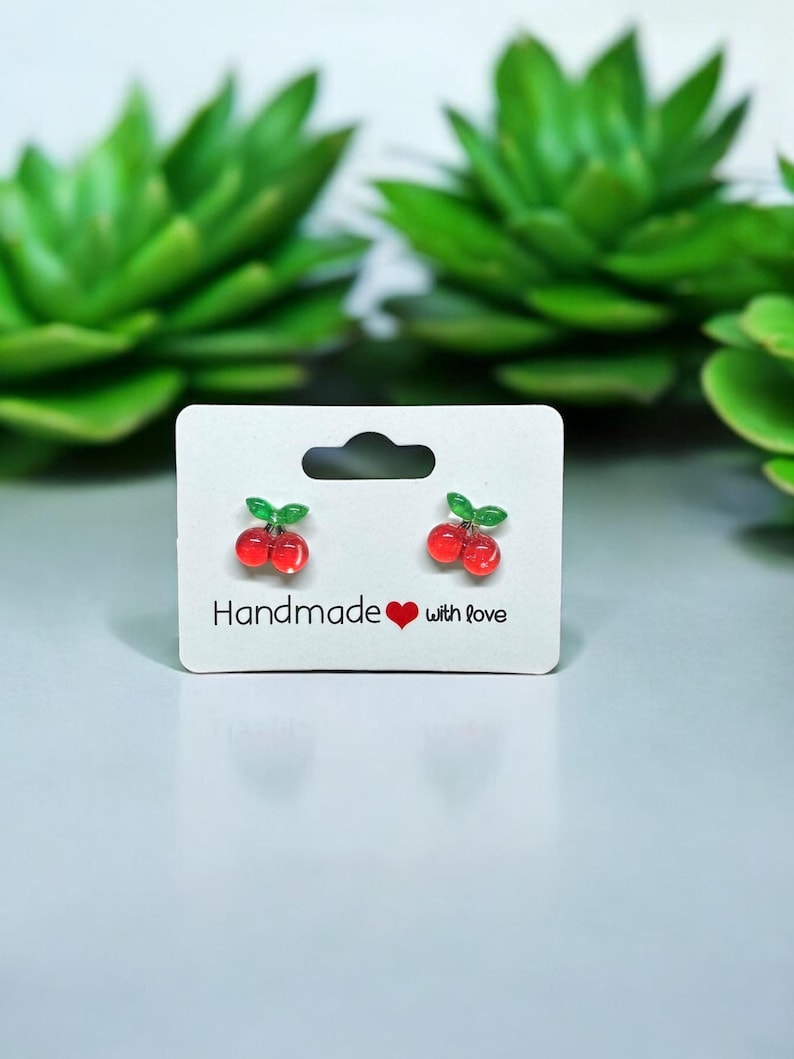 以下が含まれることがあります： 白いカードに「Handmade with Love」と書かれた、チェリーの形をしたイヤリングのペア。イヤリングは赤と緑で、光沢のある仕上げになっています。