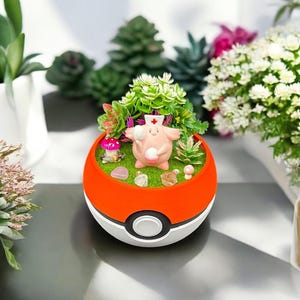 Op de afbeelding: Een decoratieve plantenbak in de vorm van een rood-witte bal, met een miniatuurlandschap met een roze stripfiguur met een verpleegstershoed. De plantenbak bevat kleine planten, stenen en een paddenstoel, wat een grillige sc&egrave;ne cre&euml;ert.