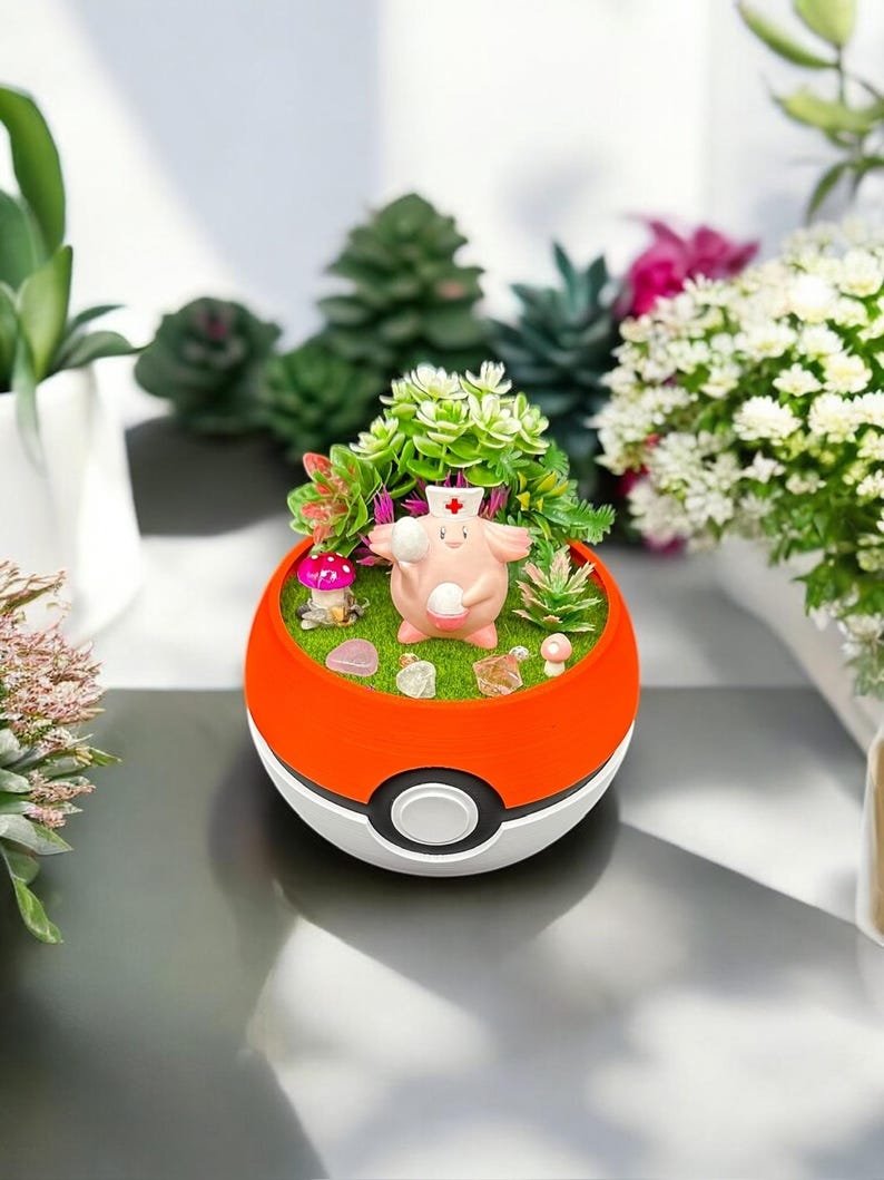 Op de afbeelding: Een decoratieve plantenbak in de vorm van een rood-witte bal, met een miniatuurlandschap met een roze stripfiguur met een verpleegstershoed. De plantenbak bevat kleine planten, stenen en een paddenstoel, wat een grillige sc&egrave;ne cre&euml;ert.