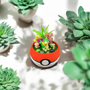 Peut inclure: Un petit pot de fleurs rond, rouge et blanc, ressemblant à une Poké Ball. Il contient un jardin miniature avec une figurine verte, de petites plantes et des accents colorés. Autour, plusieurs plantes succulentes vertes dans des pots blancs.